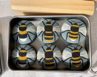 May include: Six round fabric buttons with a bee design. The bees are black with yellow stripes and are on a blue and white background. The buttons are in a metal tin.