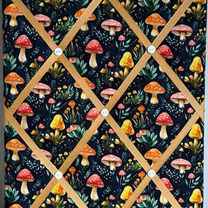 Op de afbeelding: Een stoffen memobord met een donkerblauwe achtergrond bedekt met kleurrijke paddenstoelillustraties. Het bord heeft een raster van gouden linten die op hun plaats worden gehouden door witte knopen en een bijpassende lintlus om op te hangen.