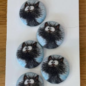 May include: Five circular fabric-covered buttons arranged on a white tag. Each button features a printed illustration of a fluffy black cat with wide white eyes and a light blue background. The buttons are round with a soft, textured fabric surface.