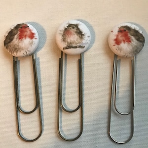 Puede incluir: Tres clips de papel metálicos con tapas redondas cubiertas de tela. Cada tapa presenta una pintura de acuarela de un petirrojo con detalles rojos, marrones y blancos. Los clips de papel están dispuestos verticalmente sobre un fondo blanco.