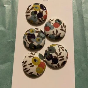 Könnte beinhalten: Fünf stoffbezogene Knöpfe mit einem Blumenmuster in Blau-, Grün-, Gelb- und Rottönen auf weißem Hintergrund. Die Knöpfe sind an einem weißen Etikett mit dem Text "Buttonhome" unten befestigt.