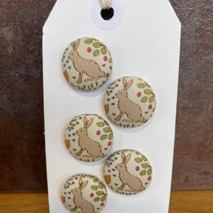 May include: A set of five fabric-covered buttons with a bunny rabbit design. The buttons are beige with a cream background and feature a rabbit, green leaves, and red berries. The buttons are attached to a white tag with the text "Buttonhome".