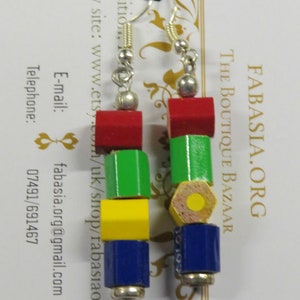 May include: A pair of earrings made from wooden pencil pieces. The earrings are red, green, yellow, and blue. The earrings are shaped like a pencil with a hexagonal barrel and a round eraser. The earrings are hanging from silver hooks.