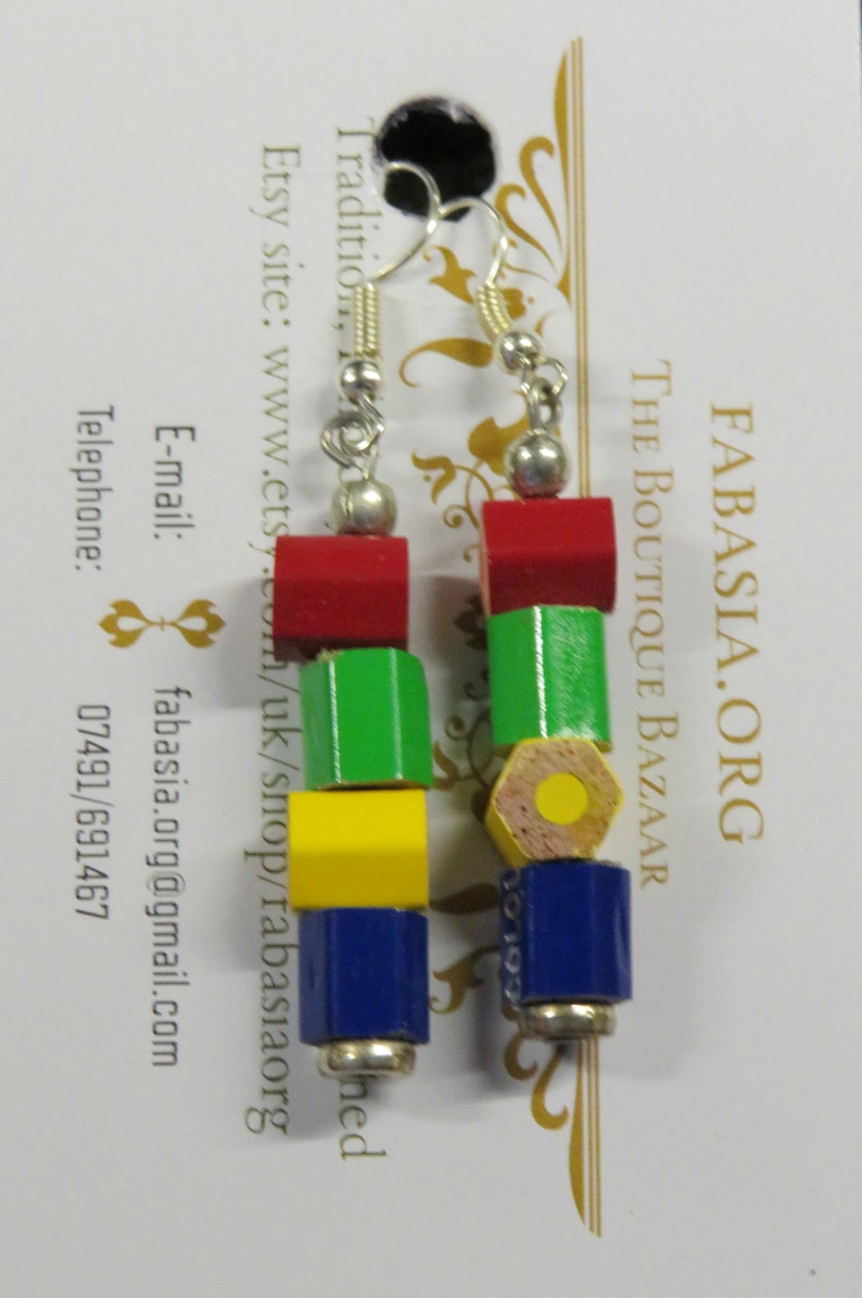 May include: A pair of earrings made from wooden pencil pieces. The earrings are red, green, yellow, and blue. The earrings are shaped like a pencil with a hexagonal barrel and a round eraser. The earrings are hanging from silver hooks.