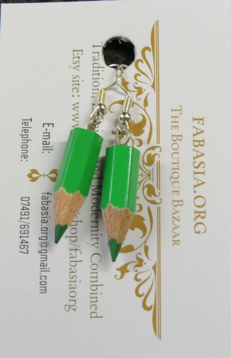 May include: A pair of green pencil earrings with silver hooks. The earrings are made to look like sharpened pencils with a wood-colored base and a green painted tip.
