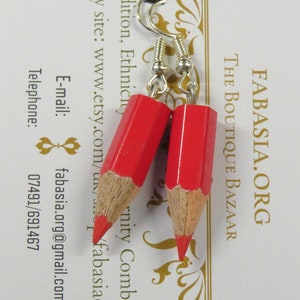 May include: A pair of red pencil earrings with silver hooks. The earrings are made from real pencils and have a wood grain finish. The earrings are hanging from a white card with the text "FABASIA.ORG THE BOUTIQUE BAZAAR Tradition, Ethnicity, Etsy site: www.etsy.com/u/fabasiaorg E-mail: fabasia.org@gmail.com Telephone: 07491/691467" printed on it.