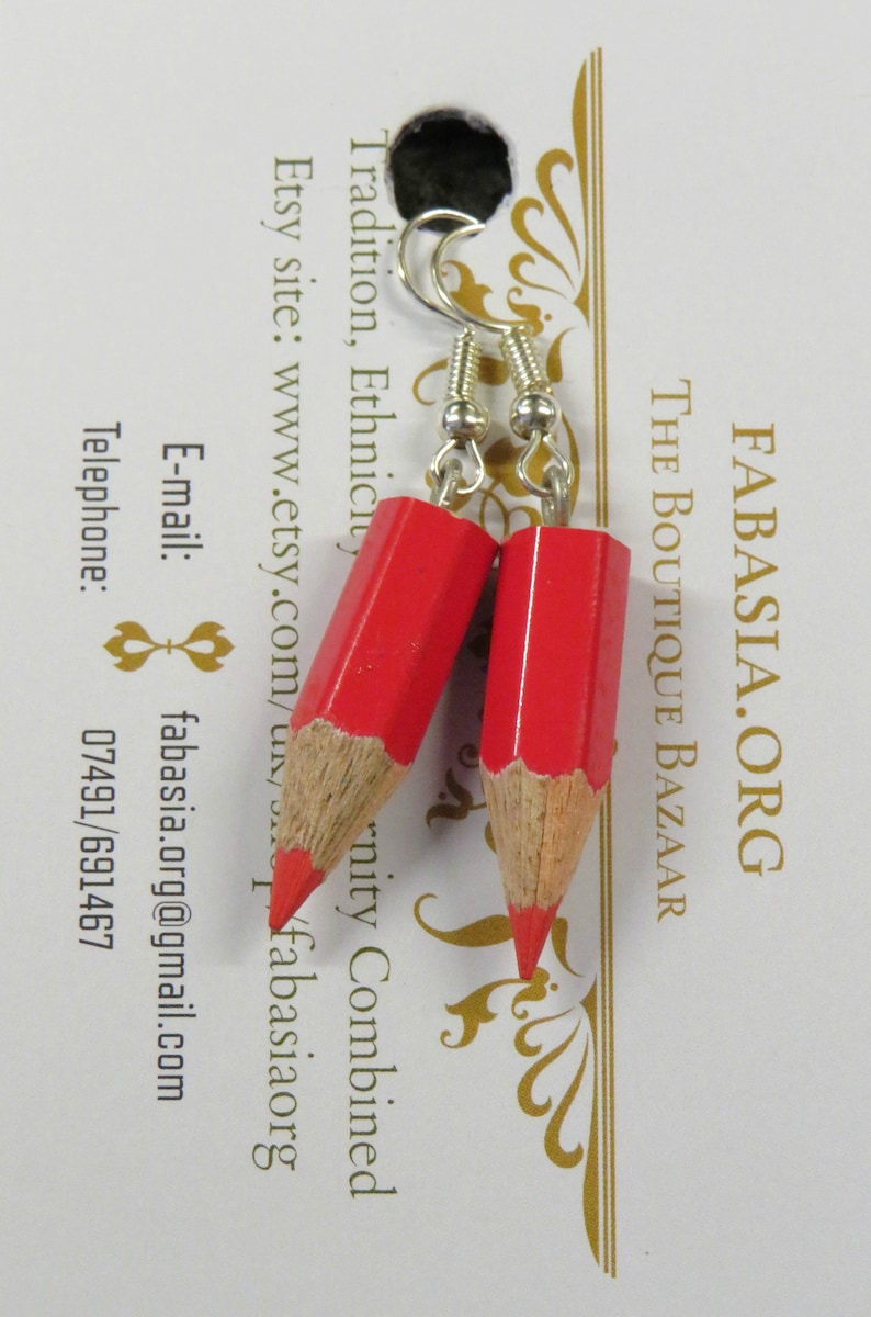 May include: A pair of red pencil earrings with silver hooks. The earrings are made from real pencils and have a wood grain finish. The earrings are hanging from a white card with the text "FABASIA.ORG THE BOUTIQUE BAZAAR Tradition, Ethnicity, Etsy site: www.etsy.com/u/fabasiaorg E-mail: fabasia.org@gmail.com Telephone: 07491/691467" printed on it.