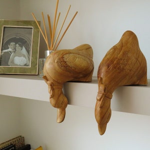 May include: Two wooden duck figurines, carved in a light brown color, are perched on a white shelf. The ducks are facing downwards, with their heads and necks extending over the edge of the shelf.