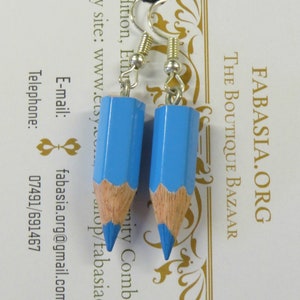 May include: A pair of blue pencil earrings with silver hooks. The pencils are about 2 inches long and have a light blue paint finish with a wood-colored tip and a blue eraser. The earrings are hanging from a white card with gold text that reads "FABASIA.ORG THE BOUTIQUE BAZAAR Tradition, EiN mity Combined Etsy site: www.etsy.com/tabasiaorg E-mail: fabasia.org@gmail.com Telephone: 07491/691467"
