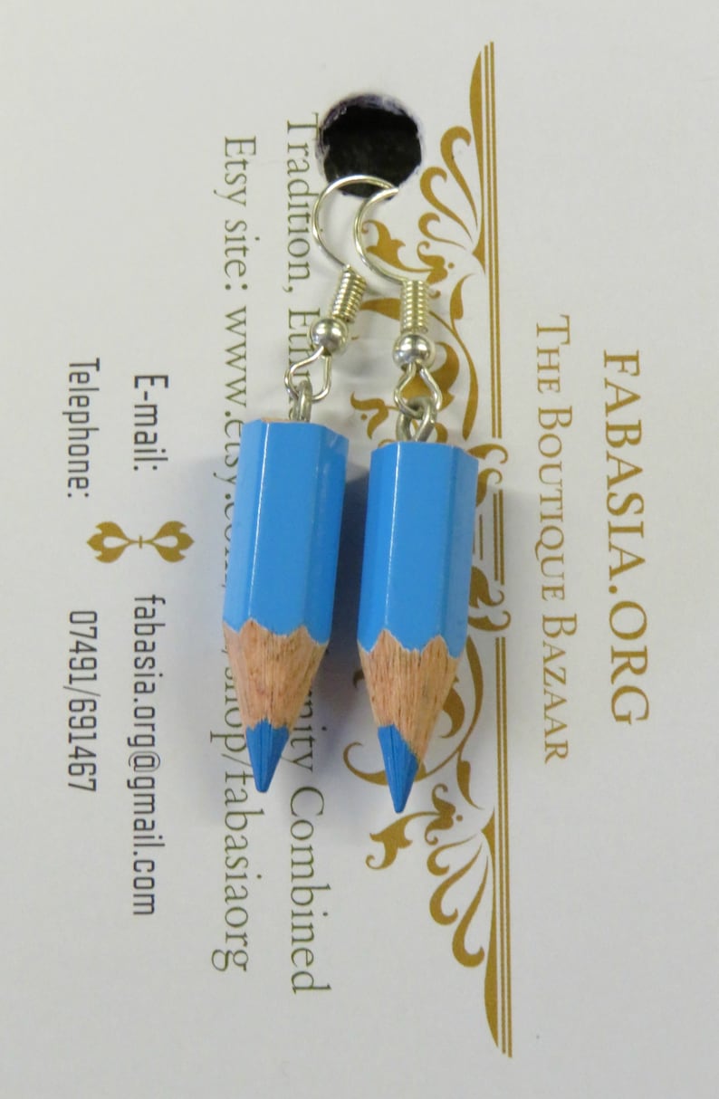 May include: A pair of blue pencil earrings with silver hooks. The pencils are about 2 inches long and have a light blue paint finish with a wood-colored tip and a blue eraser. The earrings are hanging from a white card with gold text that reads "FABASIA.ORG THE BOUTIQUE BAZAAR Tradition, EiN mity Combined Etsy site: www.etsy.com/tabasiaorg E-mail: fabasia.org@gmail.com Telephone: 07491/691467"