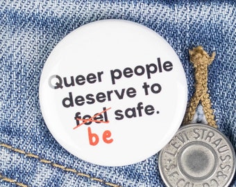 May include: A white button with black text that reads "Queer people deserve to feel safe." The word "be" is crossed out with a red line. The button is pinned to a blue denim jacket.