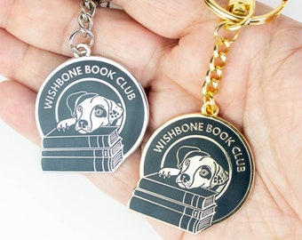 Puede incluir: Dos llaveros negros y dorados con un diseño de perro y libros. Los llaveros tienen el texto "Wishbone Book Club".