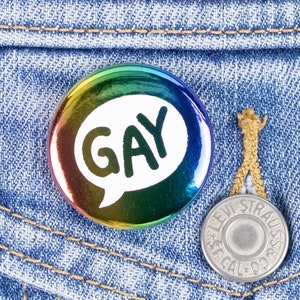 Puede incluir: Un botón redondo con un degradado arcoíris y la palabra "GAY" en un bocadillo está prendido en un bolsillo de una chaqueta vaquera. También se ve un botón plateado de Levi Strauss.