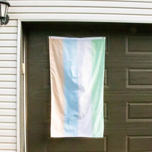 Peut inclure: Un drapeau multicolore à cinq bandes verticales. Les bandes sont de couleur pêche, bleu clair, blanc, vert clair et vert foncé. Le drapeau est accroché à une porte de garage brune.