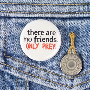 May include: A white button with the text "there are no friends. ONLY PREY." in black and red lettering. The button is pinned to a blue denim jacket.