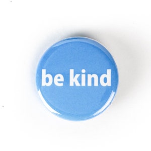 Puede incluir: Un botón redondo azul con el texto "be kind" en blanco.