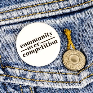 May include: A white button with black text that reads "community over competition" pinned to a blue denim jacket.