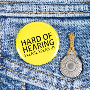 Può includere: Un bottone giallo con testo nero che recita "Hard of Hearing Please Speak Up" appuntato su una giacca di jeans blu.