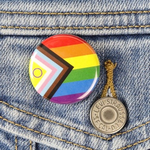 May include: A rainbow pride pin with a brown, pink, light blue, yellow, green, light blue, and purple stripe pattern. The pin is attached to a denim jacket with a Levi Strauss & Co. button.