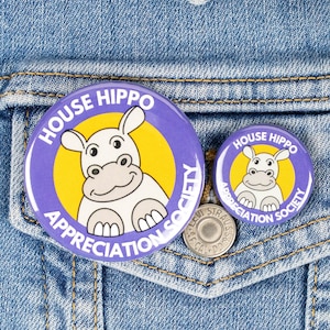 May include: Two purple and yellow House Hippo Appreciation Society buttons featuring a cartoon hippopotamus, pinned to a denim jacket.