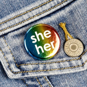 Puede incluir: Un botón de color arcoíris con las palabras "she her" en texto blanco, sobre una chaqueta vaquera. También es visible un botón plateado de Levi Strauss & Co. El denim tiene costuras amarillas.