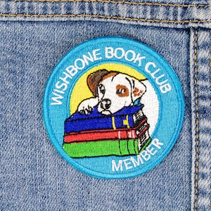 May include: A blue and white embroidered patch featuring a dog and books. The text on the patch reads "Wishbone Book Club Member".