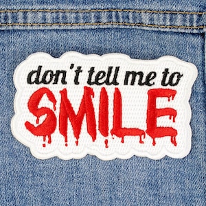 Puede incluir: Un parche blanco con una fuente roja que gotea que dice "no me digas que SONRÍA" está cosido en una chaqueta vaquera azul.