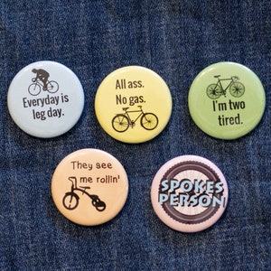 May include: Five round buttons with different cycling-themed designs. The buttons are yellow, green, blue, pink, and orange. The text on the buttons reads: "All ass. No gas." "Everyday is leg day." "I'm two tired." "They see me rollin'" "SPOKES PERSON"