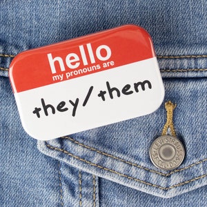 Puede incluir: Un botón rojo y blanco con el texto "hello my pronouns are they/them" en una chaqueta vaquera azul.
