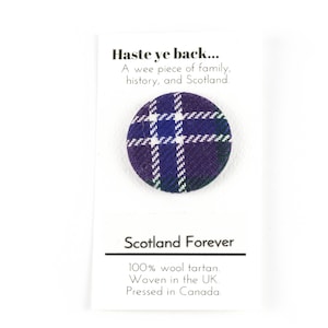 May include: A small round button with a purple, blue, white, and green tartan pattern. The button is on a white card with the text "Haste ye back... A wee piece of family, history, and Scotland. Scotland Forever 100% wool tartan. Woven in the UK. Pressed in Canada."