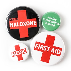 May include: Four round buttons with red crosses and white text. The buttons read "I carry naloxone", "First Aid", "Medic", and "Suicide Intervention Trained".