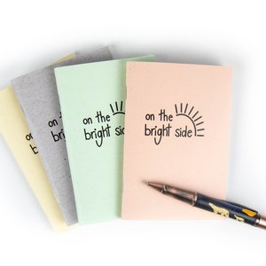 May include: Four small notebooks with different colored covers, each with the text "on the bright side" and a sun graphic. A pen with a black and brown design is lying on top of the notebooks.