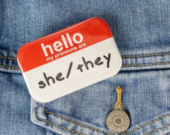 May include: A red and white button with the text "hello my pronouns are she/they" on a blue denim jacket.