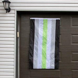 Op de afbeelding: Een groen-wit-zwarte gestreepte vlag die aan een bruine garagedeur hangt. De vlag heeft een enkele groene streep in het midden.