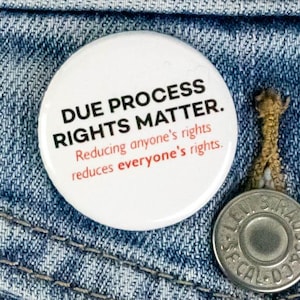 Pode incluir: Um botão branco com o texto "DUE PROCESS RIGHTS MATTER." está preso no bolso de uma jaqueta jeans azul. O texto "Reducing anyone's rights reduces everyone's rights" também está presente.