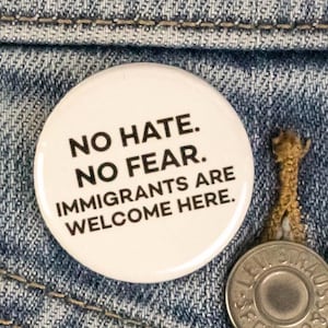 May include: A white button with the text "NO HATE. NO FEAR. IMMIGRANTS ARE WELCOME HERE." is pinned on a blue denim jacket. A silver button is visible near the button.