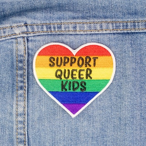 May include: A rainbow heart-shaped patch with the text "SUPPORT QUEER KIDS" on a denim background.