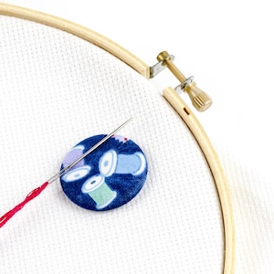 May include: A close-up of a wooden embroidery hoop with a white fabric stretched across it. A red thread is threaded through a needle and a blue button with a colorful spool design is attached to the fabric.