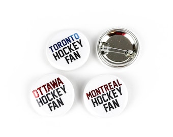 Op de afbeelding: Vier witte ronde knoppen met zwarte tekst en verschillende kleuraccenten. De knoppen tonen "Toronto Hockey Fan" in blauw, "Ottawa Hockey Fan" in rood, "Montreal Hockey Fan" in donkerrood, en de vierde knop is de achterkant van een knop met een zilveren pin.