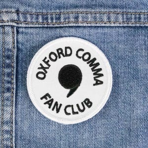 May include: A white circular patch with black text that reads "Oxford Comma Fan Club" and a black comma symbol in the centre. The patch is sewn onto a blue denim jacket.