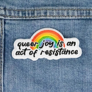 Könnte beinhalten: Ein weißer Patch mit einem Regenbogenbogen und dem Text "queer joy is an act of resistance" auf einer blauen Jeansjacke.