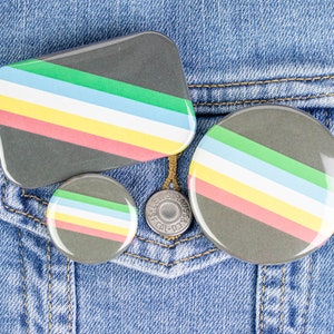 May include: Three striped badges with a black background and colourful stripes in green, blue, yellow, and pink. The badges are on a blue denim jacket.