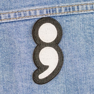 Pode incluir: Um remendo de ponto e vírgula bordado em preto e branco. O ponto e vírgula é um símbolo de esperança e força.