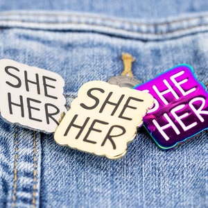 Peut inclure: Trois épingles en émail sur une veste en jean bleue. Les épingles sont toutes carrées et ont des coins arrondis. Les épingles disent "She Her" en texte noir sur un fond blanc, or et iridescent.