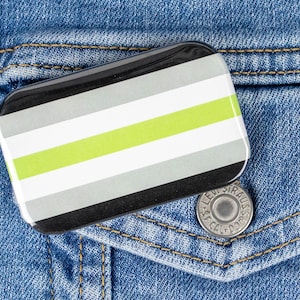 May include: A rectangular pin with a black border and a striped design in black, grey, white, and lime green. The pin is on a blue denim jacket pocket, next to a silver button.