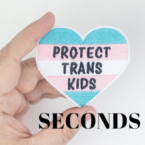 May include: A heart-shaped embroidered patch featuring horizontal stripes in light blue, pink, and white. The text "PROTECT TRANS KIDS" is stitched in dark blue thread across the centre of the patch, which has a white border.