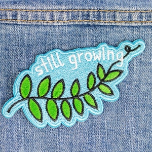 Puede incluir: Parche bordado con la frase "still growing" en blanco, sobre un diseño de hojas verdes con contornos negros. El parche tiene un fondo azul claro y un borde azul, sobre un fondo de mezclilla.