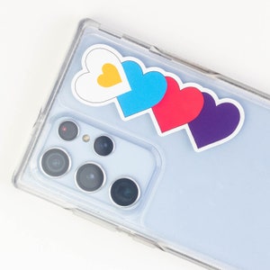 Puede incluir: Una funda transparente para teléfono con cuatro pegatinas en forma de corazón. Las pegatinas están superpuestas y son de color blanco, azul, rojo y morado.