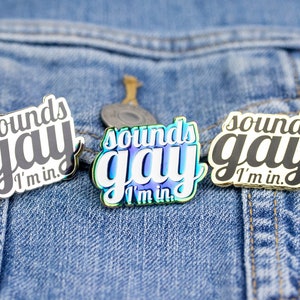 Peut inclure: Trois épingles en émail avec le texte "Sounds Gay I'm in." Les épingles sont sur une veste en jean bleue. L'épingle du milieu a une finition arc-en-ciel irisée.