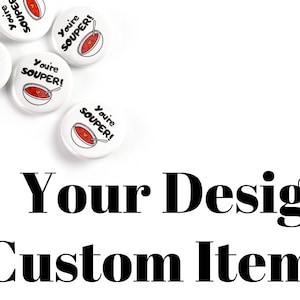 May include: White round buttons with the text "You're Souper!" and a red bowl of soup with a spoon. The buttons are scattered on a white surface. The text "Your Design Custom Items" is below the buttons.